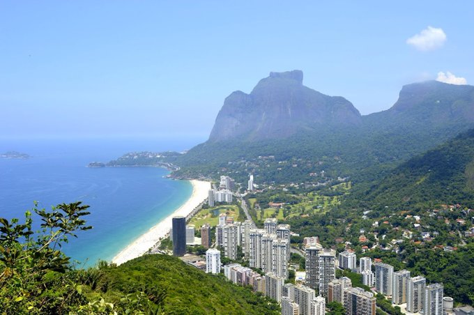 Aerial view of Rio de Janeiro showing the coastline, beach, high-rise buildings, and the iconic mountains in the background.