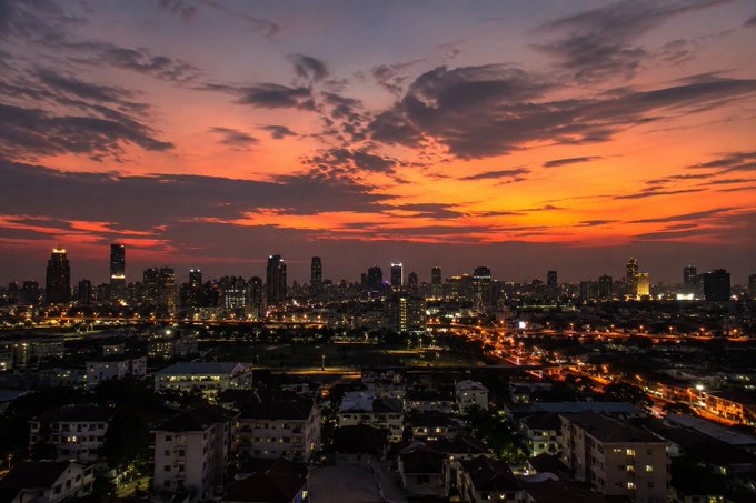 A vibrant city skyline at sunset, with warm orange and pink hues illuminating the sky above silhouetted skyscrapers and city lights.