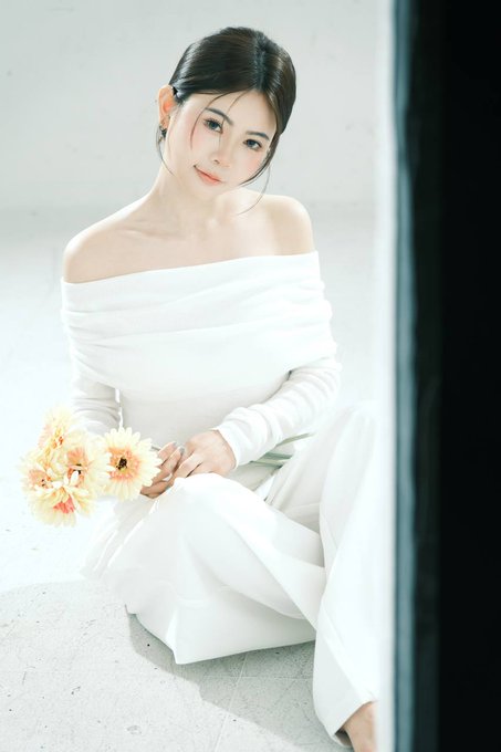 A portrait of a young woman with dark hair wearing a white off-the-shoulder long-sleeved dress, sitting on a light-colored floor and holding a small bouquet of pale flowers.