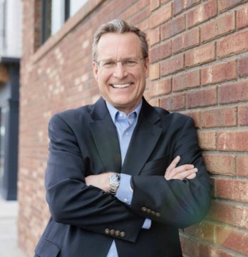 A smiling older man in a dark blazer stands with arms crossed against a red brick building.