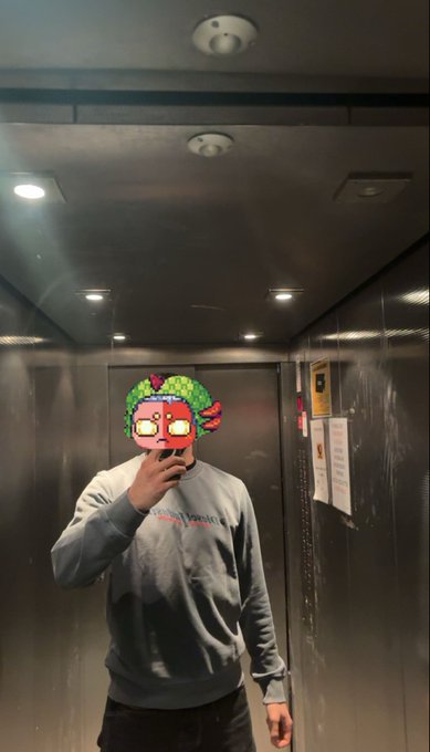 The image shows a man taking a selfie inside an elevator, wearing a gray sweatshirt with a green design on the front. The background of the photo features a metal ceiling with lights that are turned off, giving the impression that the photo was taken in a dimly lit space or during nighttime hours. There is also a postcard-like sign visible in the image, which could be related to the man's selfie or another event taking place within the elevator.