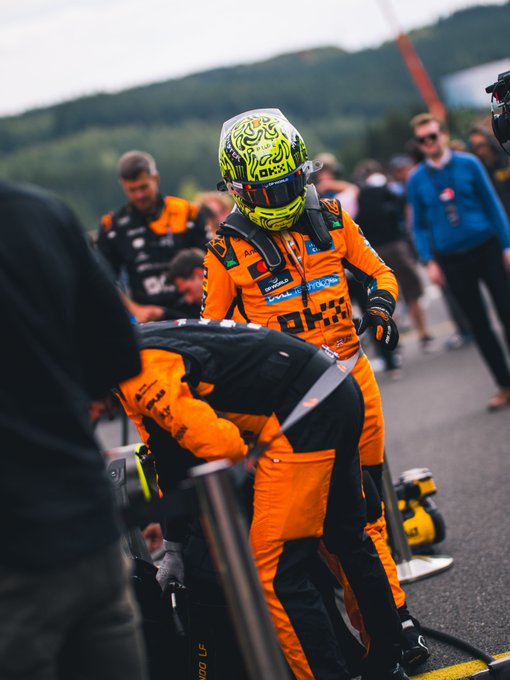 An F1 driver in a bright orange racing suit and a neon yellow helmet stands in the pit lane next to a crew member, preparing to make adjustments.