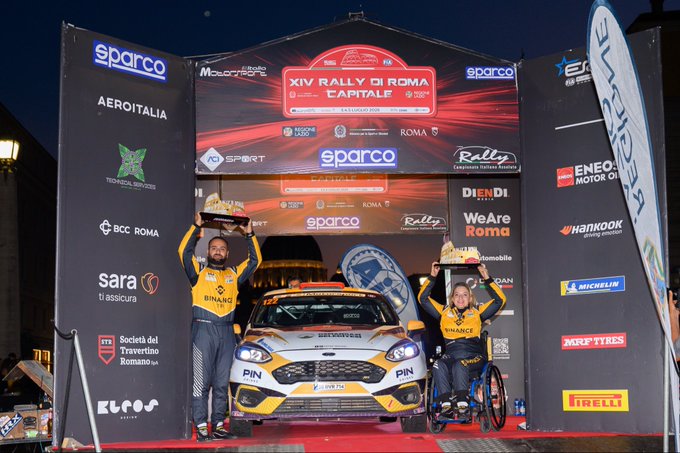 Two rally drivers, one in a wheelchair, celebrate at the "XIV Rally di Roma Capitale" with trophies and a rally car. This image showcases the triumph of determination and belief, as highlighted by Kübra Denizci Keskin's accomplishments.