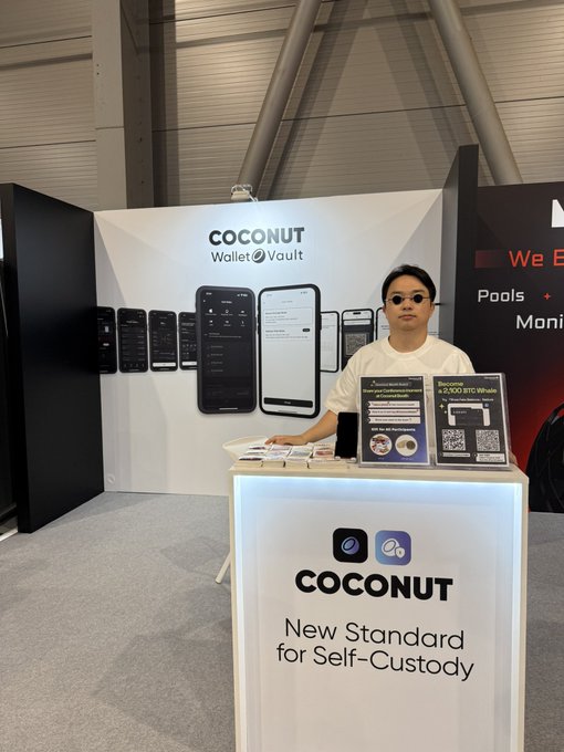 The image features a man standing behind a table with a sign that reads "Coconut Wallet". The man appears to be wearing sunglasses, giving him an air of mystery or intrigue. He is positioned next to a display case filled with various items, including cell phones and other accessories. The background shows a large room with a white ceiling and walls, which provides ample space for the event taking place in this area.