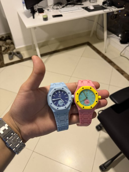 A hand displays two pastel-colored wristwatches (light blue and pink with yellow accents) against a tiled floor and desk background. The scene reflects a personal, lifestyle-oriented post tagged with $LARP, suggesting the watches or the moment are being shared in a relaxed, perhaps playful context.