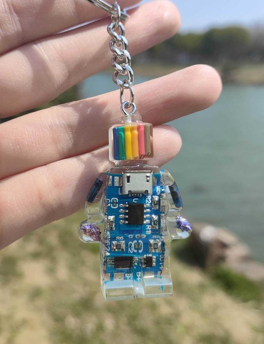 A hand holds up a clear resin LEGO minifigure keychain that encapsulates a blue printed circuit board (PCB) with electronic components and colorful resistor bands, set against a blurred background of water and grass.