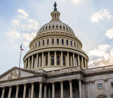 The image shows the iconic dome of the U.S. Capitol Building with an American flag visible against a cloudy sky, symbolizing the seat of the U.S. Senate where, as House Majority Whip Tom Emmer notes, the decision on the CLARITY Act now rests.