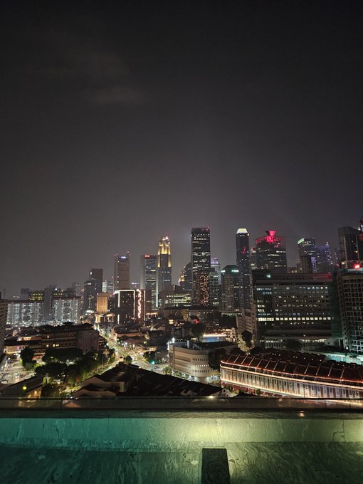 A nighttime cityscape view from a rooftop or high vantage point shows illuminated skyscrapers, city lights, and what appears to be a swimming pool edge in the foreground.
