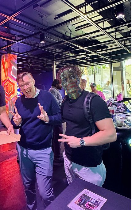 The image shows two men posing together with one of them wearing a mask that resembles a skeleton or a monster face. They are standing next to each other at an event, possibly a signing event for a book. The man without the mask has his hand in the air and is smiling, while the person behind him also has their hand raised in the air. In the background, there are several people present, indicating that it might be a social gathering or a public event.