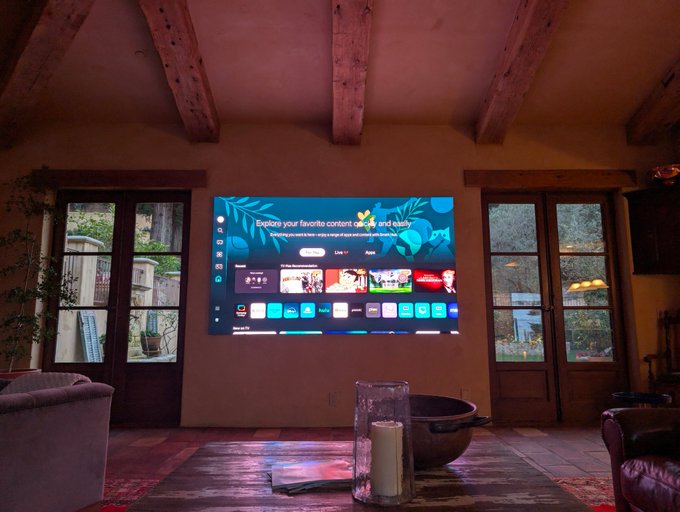 A cozy living room with exposed wooden beams features a large wall-mounted smart TV displaying a colorful home screen with the headline "Explore your favorite content casually and easily," flanked by French doors leading to an outdoor patio. The scene relates to David's message by showcasing a comfortable, tech-integrated home environment that embodies effortless access to entertainment at home.