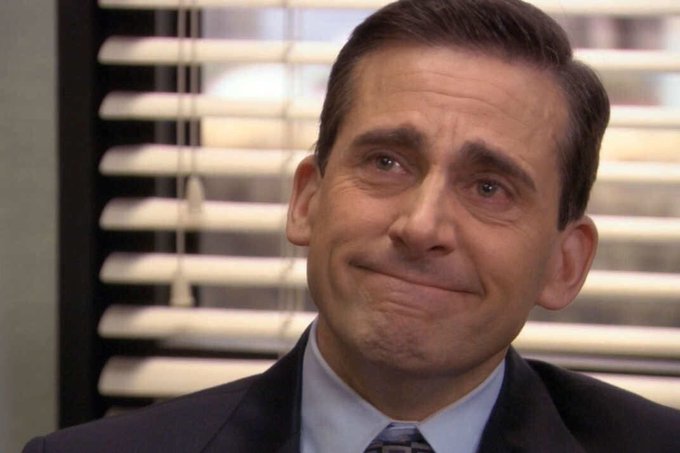 A close-up shot of Steve Carell as Michael Scott from The Office, displaying a tight-lipped, awkward smile in front of window blinds.