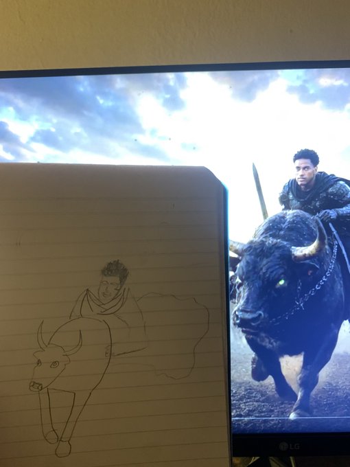 A hand-drawn sketch on lined notebook paper depicts a person riding a bull, placed next to an LG monitor showing a similar scene of a warrior riding a large black bull against a cloudy sky.