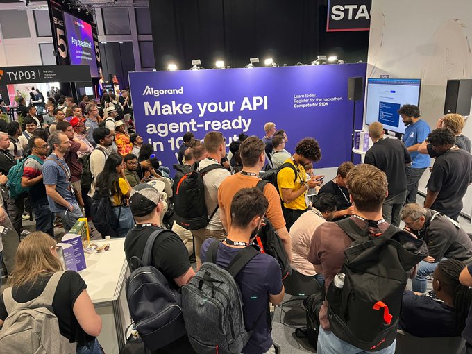 A crowded tech conference floor where attendees gather around an Algorand booth with signage reading 'Make your API agent-ready in 10' and 'Learn how Register for the hackathon Compete for $10K'.