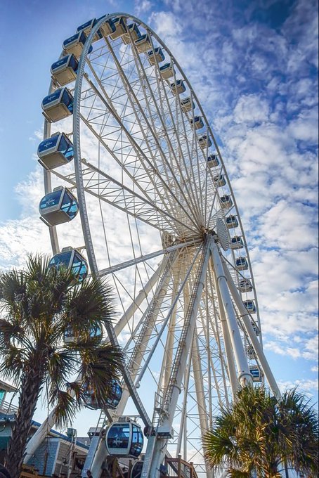 A low-angle photograph of a large white Ferris wheel against a partly cloudy blue sky, with palm trees visible at the bottom.
