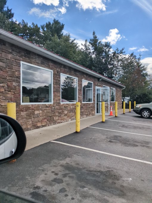 A small commercial building with stacked stone exterior walls, white-framed windows, and a glass door sits behind a parking lot lined with yellow bollards.
