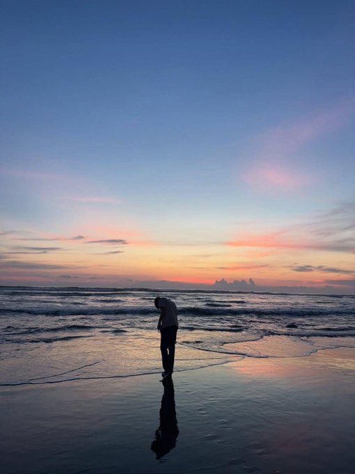 A silhouetted figure stands on a wet beach at sunset, with their reflection visible on the smooth sand and a colorful pink and orange sky above.