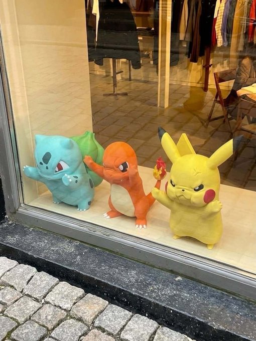 Three Pokémon figurines—Bulbasaur, Charmander, and Pikachu—stand on a ledge outside a shop window with clothing displays visible behind the glass.