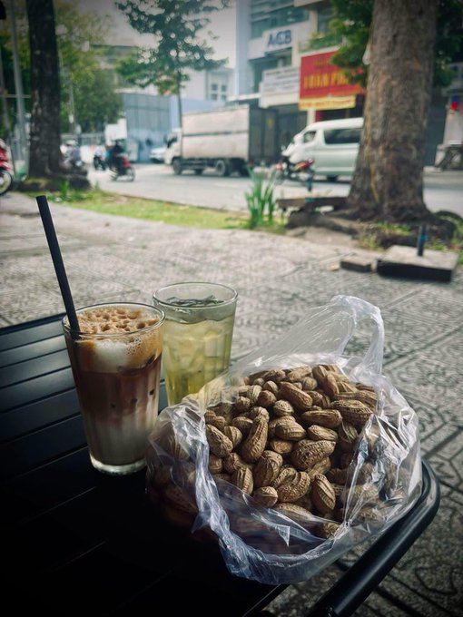 An iced coffee and iced tea alongside a plastic bag of unshelled peanuts on a black chair, with a street scene visible in the background.