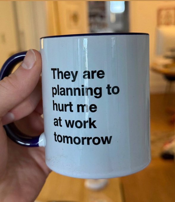 A hand holds a white mug with blue trim that reads: "They are planning to hurt me at work tomorrow".