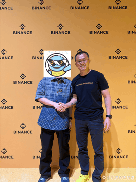 Two men pose together at a Binance event, with one wearing a duck face emoji mask over their head, in front of a yellow backdrop featuring the Binance logo.