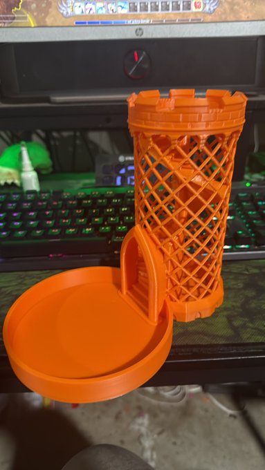 An orange 3D-printed cylindrical object resembling a miniature tower or castle, displayed on a desk in front of a glowing RGB keyboard and an HP monitor.
