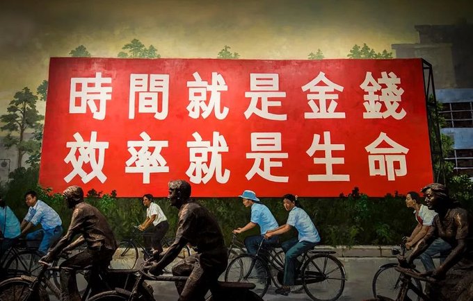 A sculptural scene depicting cyclists and figures riding bicycles past a large red billboard with white Chinese characters reading "時間就是金錢 效率就是生命" (Time is money, efficiency is life).
