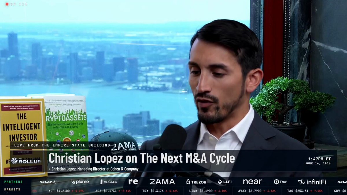 The image features a man wearing a suit, sitting at a desk with a microphone in front of him. The background shows an office setting with a window overlooking a cityscape. There are several books placed around the room, including one titled "The Intelligent Investor" and another called "The Next M&A Cycle". These books suggest that the man might be discussing investment strategies or market trends related to the next M&A cycle. The image also includes text that reads "Christian Lopez on The Next M&A Cycle", which further supports the idea of a discussion about the next M&A cycle involving Christian Lopez.