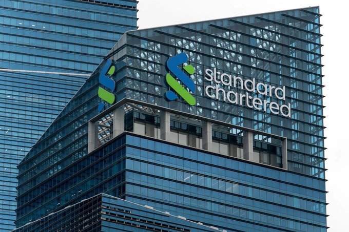 The upper floors of a modern glass skyscraper display the Standard Chartered logo and company name in blue and green lettering.