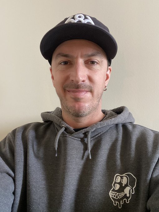 A man wearing a dark baseball cap and a gray hoodie featuring a melting smiley face graphic, smiling at the camera against a plain wall.