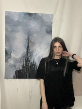 A young woman with long dark hair wearing a black shirt, standing next to her gray-toned painting of a gothic castle against a cloudy sky.
