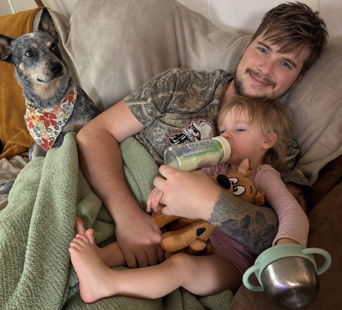 A father relaxes on a couch feeding a bottle to his toddler, who clutches a Scooby-Doo plush toy, while their gray dog watches. The scene embodies the "dad life" theme, contrasting playful market analysis (Scooby-Doo vs. YouTube TA) with the warmth of family moments.