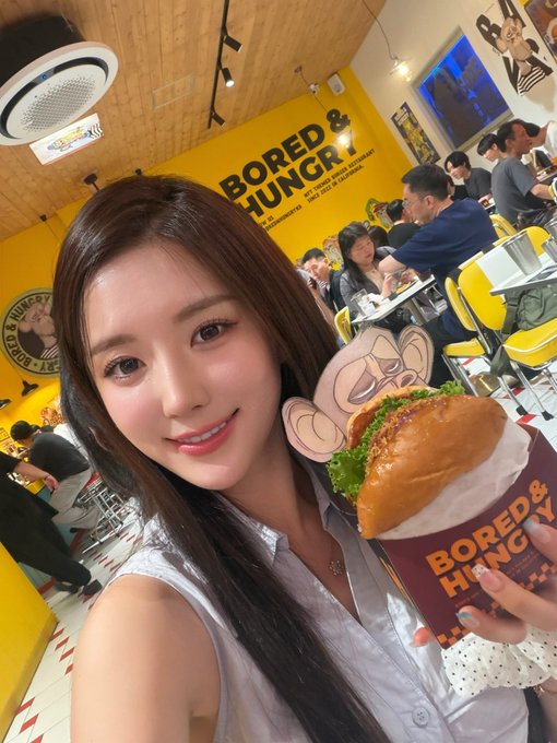 A young woman takes a selfie in a restaurant called "BORED & HUNGRY," holding a burger from the same establishment.