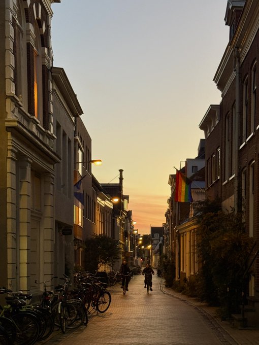 A narrow cobblestone street at sunset is lined with brick buildings, parked bicycles, and a rainbow pride flag hanging from one of the facades.