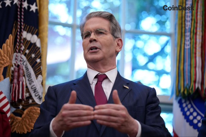 Treasury Secretary Scott Bessent, identified by the accompanying post, speaks in what appears to be a formal government setting with U.S. flags and official ribbons visible behind him. His image accompanies a statement about the Treasury's efforts to implement the GENIUS Act framework to reinforce the U.S. dollar's global reserve status and maintain America's leadership in cryptocurrency. The "CoinDesk" watermark in the upper right indicates the image's source, suggesting it was taken during a press engagement related to digital asset policy.