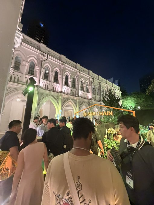 A lively nighttime gathering outside an illuminated historic building features a crowd of attendees near a glowing "BNB CHAIN" sign, capturing the vibrant social atmosphere of the BNB community coming alive after dark.