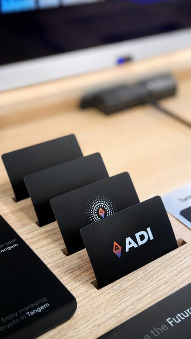 ADI-branded Tangem hardware wallet cards are displayed on a wooden retail counter, with one card showing the ADI logo and another displaying a token design, promoting the partnership's expansion into Virgin Megastores across the UAE for secure self-custody.