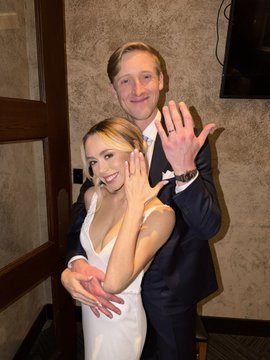 A smiling couple in formal attire pose together, both holding up their hands to display their rings in celebration of their engagement.
