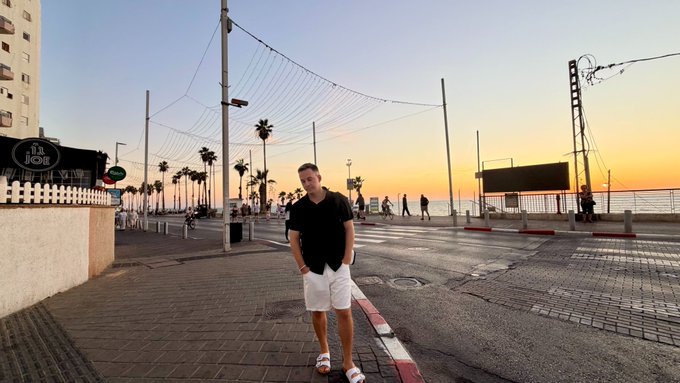 A man standing on a paved road at sunset in what appears to be a coastal or festival area, with tall light poles, palm trees, and a sunset sky in the background.