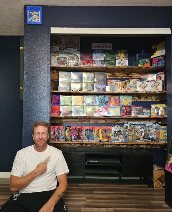 A man in a white t-shirt kneels and points upward toward a wooden shelf packed with Pokémon trading card game booster boxes and merchandise.
