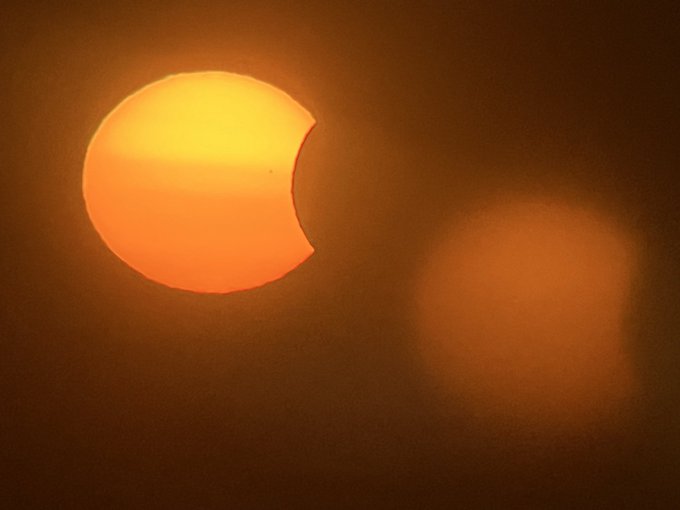 An orange-hued sun showing a partial solar eclipse, with a dark curved silhouette (the moon) biting into its upper right edge against a deep amber sky.