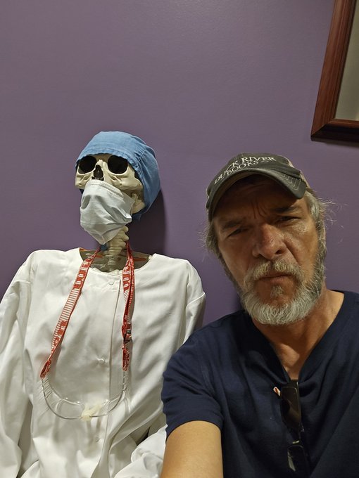 A selfie of a bearded man wearing a "BLACK RIVER" cap next to a skeleton dressed in medical scrubs, a surgical cap, and a mask.