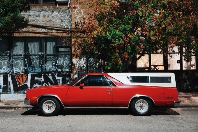 A red and white vintage coupe utility car is parked on a city street in front of a graffiti-covered brick building, with lush green vines cascading above it. The image reflects the rugged, urban aesthetic referenced in the post's call for photographs "in the style you see below," evoking a nostalgic, street-photography feel with its bold colors and gritty backdrop.