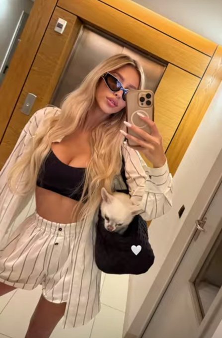 A mirror selfie of a blonde woman wearing sunglasses, a black crop top, and striped pajamas, holding a small white cat in a bag.