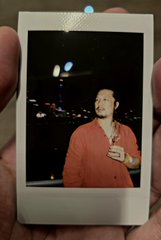 A Polaroid photo of singer Drake wearing a pinkish-orange shirt, holding it up against a dark background at night.