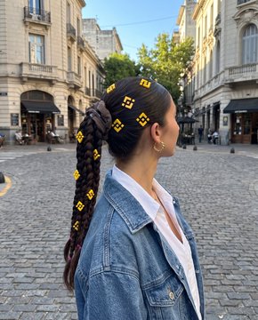 A woman with a sleek braided ponytail stands in a European-style plaza, her denim jacket and white blouse complementing the historic architecture behind her. The yellow diamond shapes scattered across the image appear to be digital embellishments highlighting her hairstyle, fitting the post's caption about "the details."