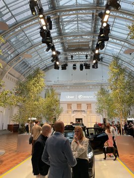 An indoor event space features a glass ceiling, hanging lights, and decorative trees, with attendees gathered around a display vehicle under a 'Liber minds' sign.