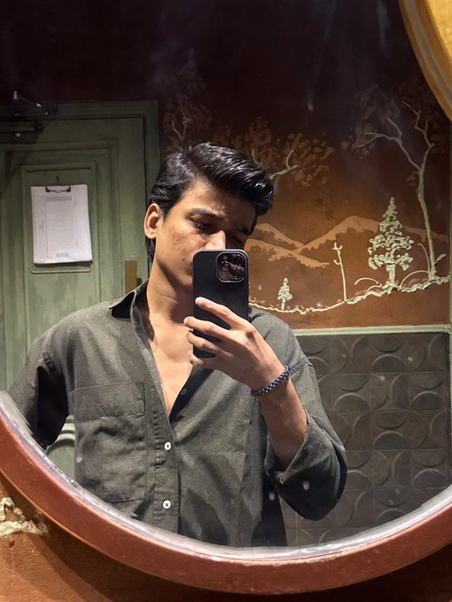 A young man with dark hair takes a mirror selfie with a smartphone in a dimly lit room featuring a mural of trees on the wall.