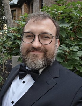 A portrait photograph shows a smiling man with a beard and glasses, dressed in a black tuxedo with a black bow tie, standing in front of green foliage.