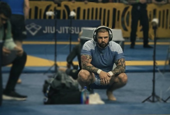 A man with tattoos and headphones squatting on a blue mat at what appears to be a Jiu-Jitsu competition or training area, looking focused.