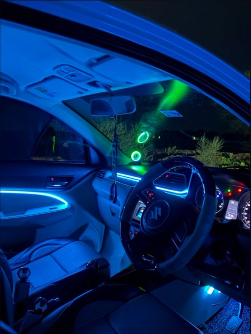 The interior of a car at night illuminated by striking blue ambient lighting along the dashboard and door panels, featuring a flat-bottomed steering wheel and green exterior lights visible through the windshield.